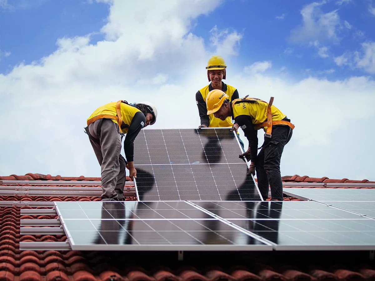 Three installers in hard hats and safety harnesses lifting a solar panel into place on a tiled roof